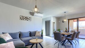 Ein modernes Wohnzimmer mit grauem Sofa, einem grünen Sessel, einem Holztisch und einem Essbereich mit Tisch und Stühlen.