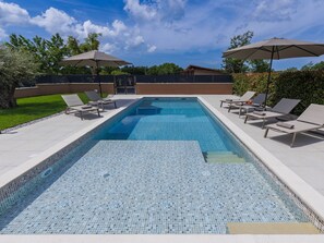 A rectangular swimming pool with tiled edges, surrounded by lounge chairs and umbrellas, set against a backdrop of trees and a clear sky. - Apartment Emili by Interhome (Vižinada)