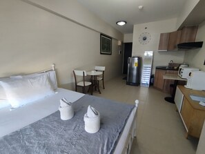 A hotel room with a bed, a small table with chairs, a refrigerator, a microwave, and a kitchen area with cabinets and a sink.