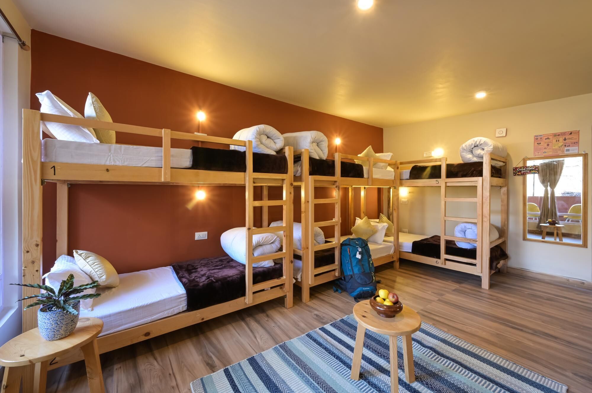 Quarto tipo dormitório com beliches, uma mesinha com uma tigela de frutas, um vaso de planta e um tapete listrado.