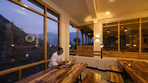 A person seated at a wooden table with a laptop, large windows with a view of a mountainous landscape, and a wooden balcony with hanging lights. - Zostel Dobhi (Kullu)