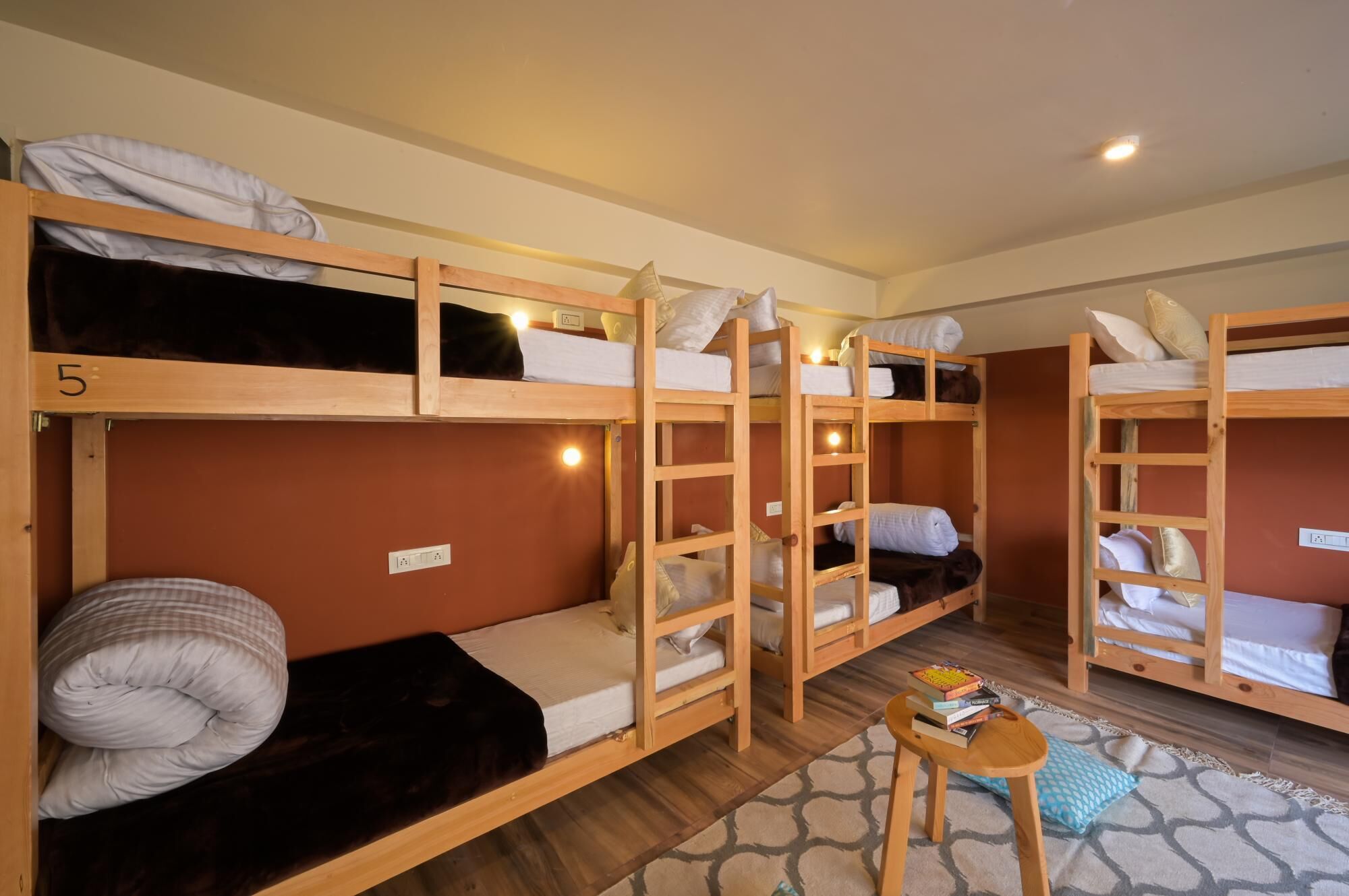 A dormitory room with bunk beds, a small table with books, and a patterned rug.