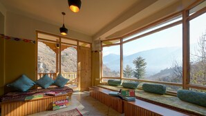 A cozy room with a large window, wooden benches, and a colorful rug. - Zostel Dobhi (Kullu)