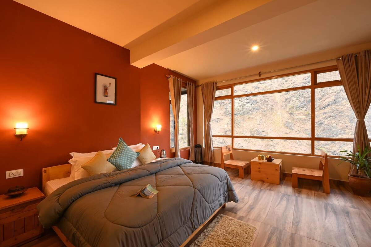 A bedroom with a large bed, two bedside tables, a chair, a small table, a potted plant, and a window with curtains.