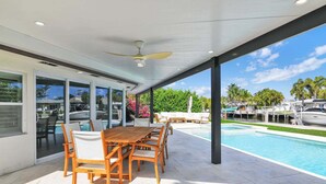 En moderne udendørs terrasse med pool, træbord og stole samt en ventilator i loftet.