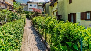 Un étroit sentier pavé, bordé d’une végétation luxuriante et de bâtiments traditionnels dotés de balcons, qui mène vers un paysage montagneux en toile de fond.