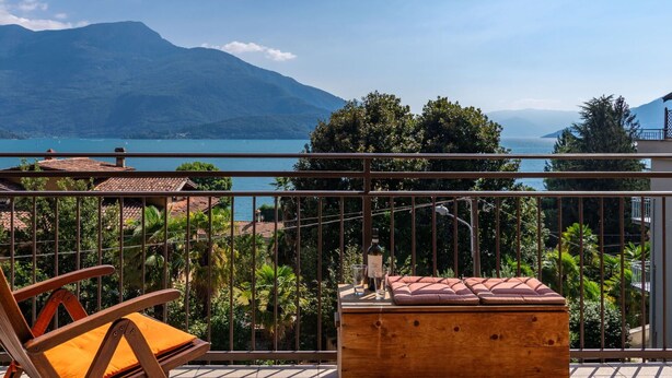 A balcony with a wooden table, a cushioned bench, and a view of a lake and mountains.