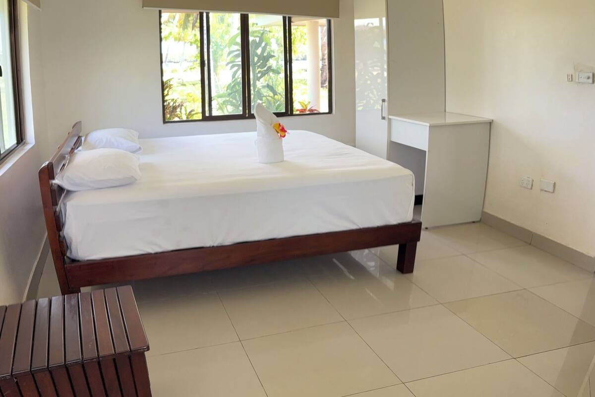 A bedroom with a large bed, a ceiling fan, and a window with a view of greenery.