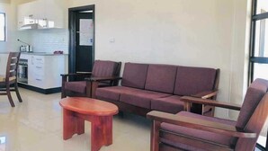 Una sala de estar con un sofá, dos sillones y una mesa de centro. Se ve un área de cocina con armarios y una estufa a través de una puerta abierta.
