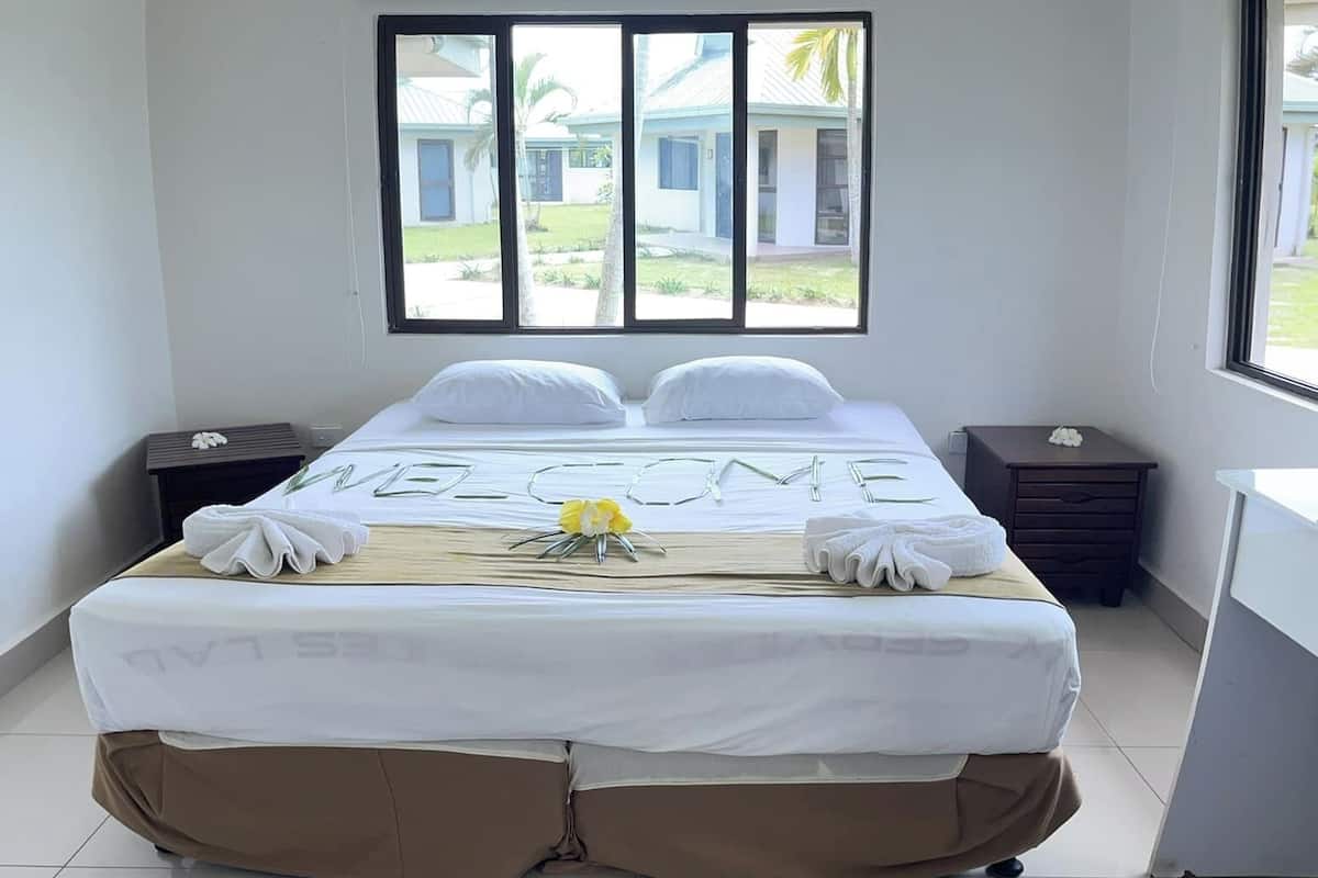 A hotel room with a large bed, two bedside tables, a desk, and a window with a view of buildings and greenery.