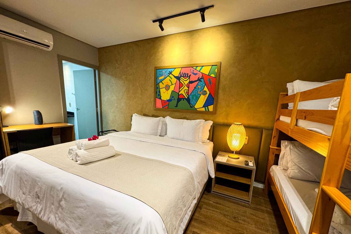 A hotel room with a large bed, a bedside lamp, a bunk bed, and a colorful abstract painting on the wall.