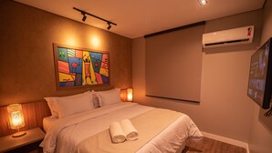 Une chambre d’hôtel avec un lit, un tableau au mur, un climatiseur et un téléviseur fixé au mur.