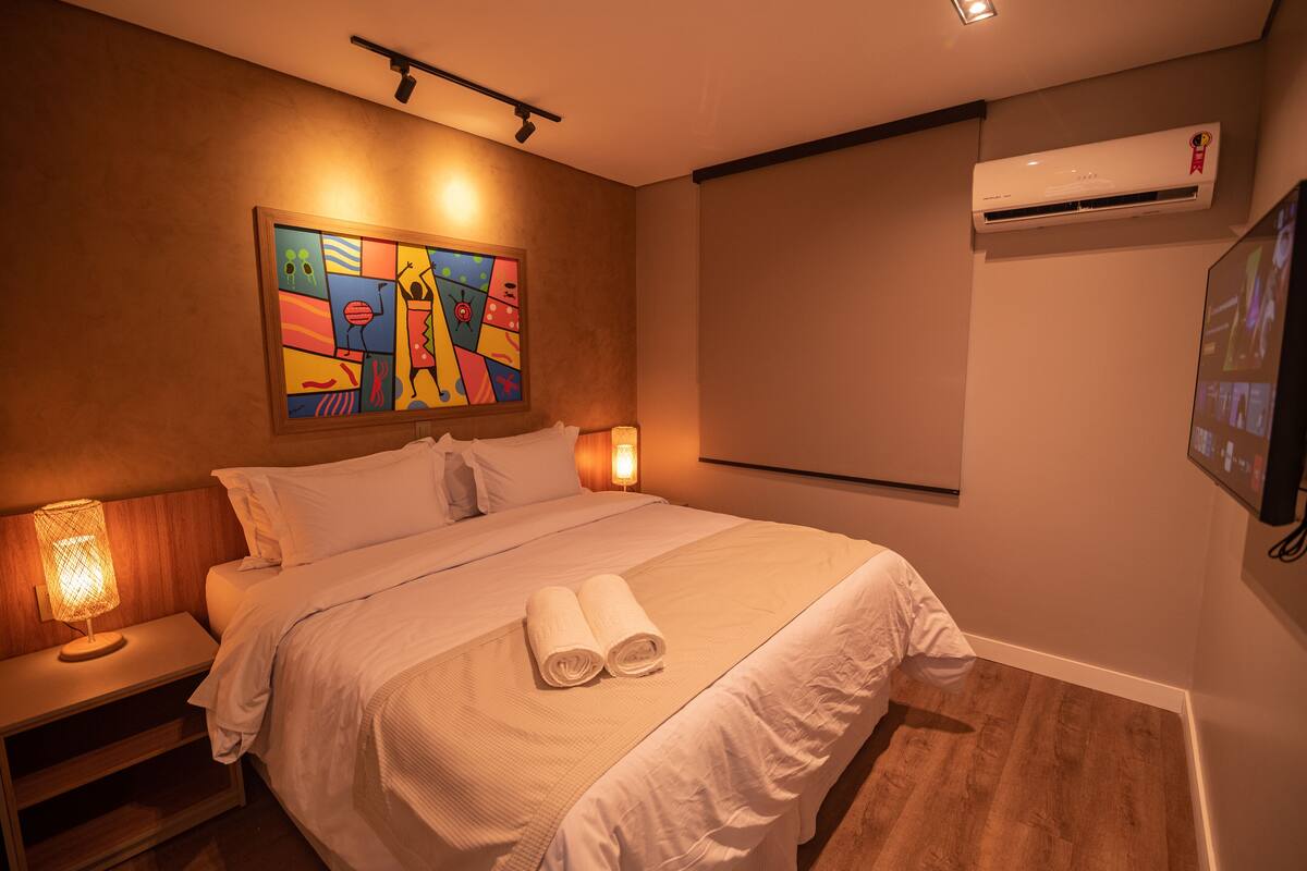A hotel room with a bed, a painting on the wall, an air conditioner, and a TV mounted on the wall.