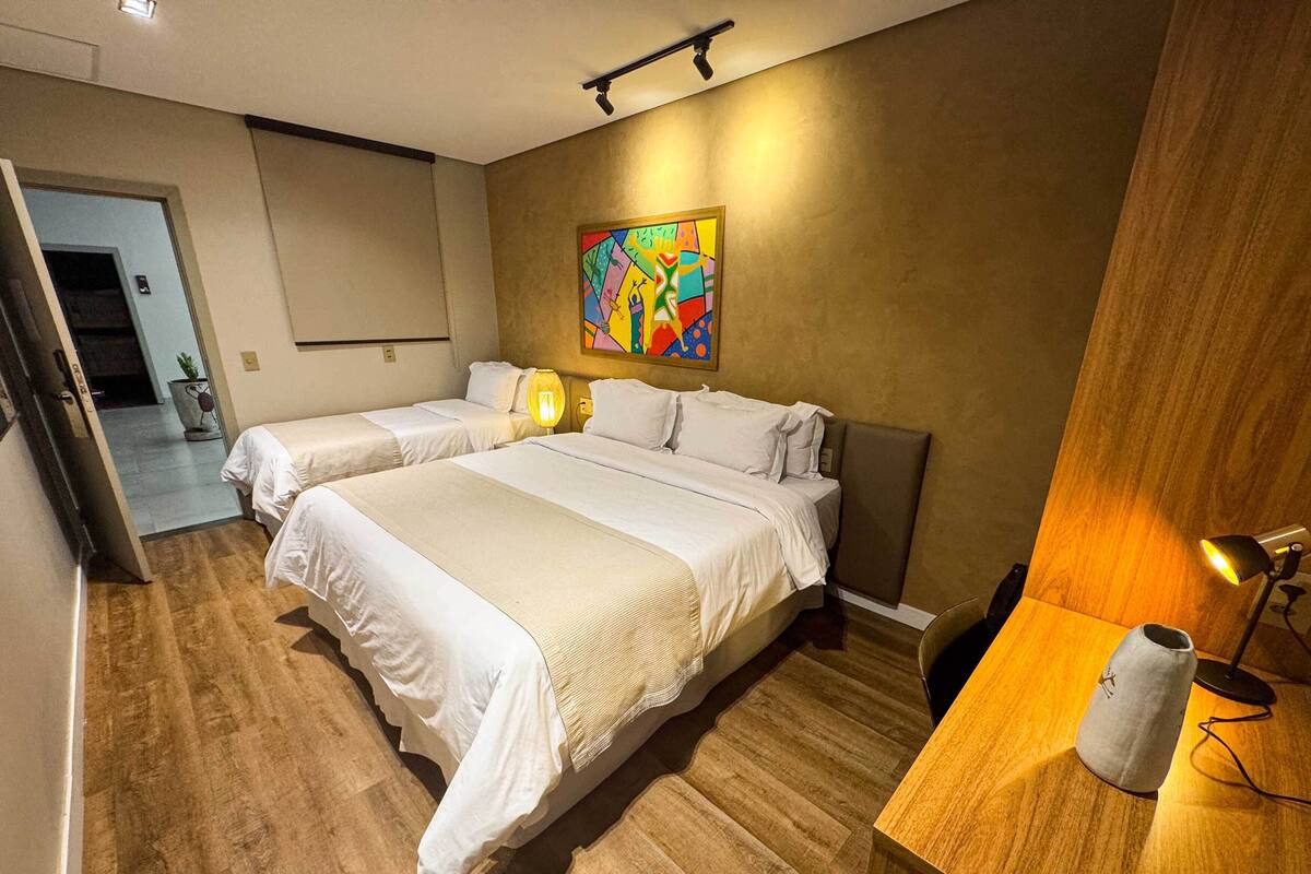 A hotel room with two beds, a wooden floor, a colorful abstract painting on the wall, a desk with a lamp, and a small plant.
