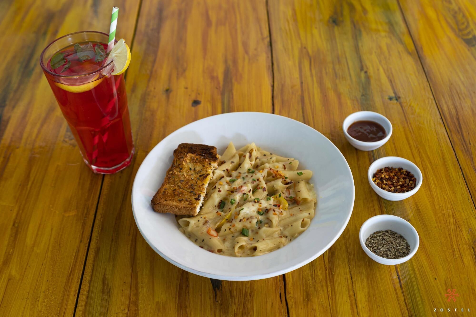 A plate of pasta with a slice of bread, a glass of red drink, and small bowls of condiments on a wooden table.
