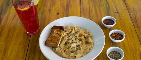 A plate of pasta with a slice of bread, a glass of red drink, and small bowls of condiments on a wooden table.