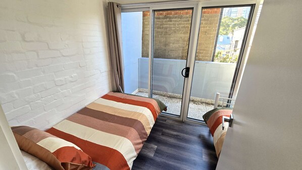 A small, narrow room with a bed, a sliding glass door leading to an outdoor area, and a white brick wall.