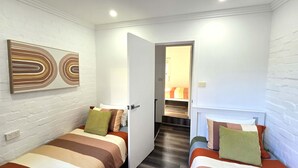 A small, modern room with two beds, a white brick wall, and a hallway leading to another room.