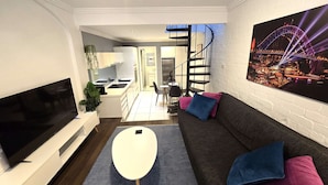 A modern living room with a flat-screen TV, a sofa with cushions, a coffee table, and a dining area with chairs and a table.