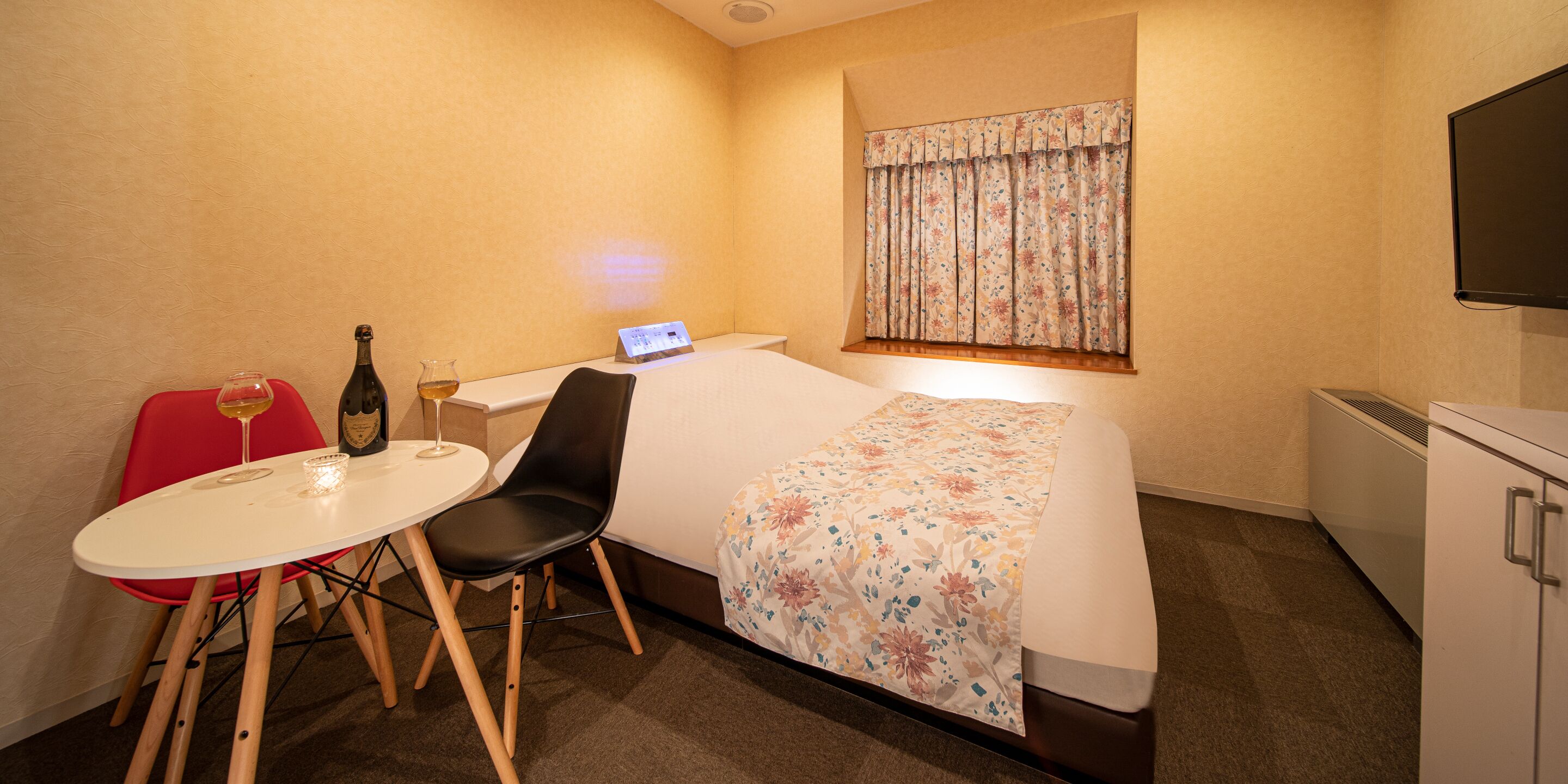 Quarto de hotel com cama, secretária com computador, cadeira, mesa pequena com uma taça e garrafa de vinho, e televisão na parede.