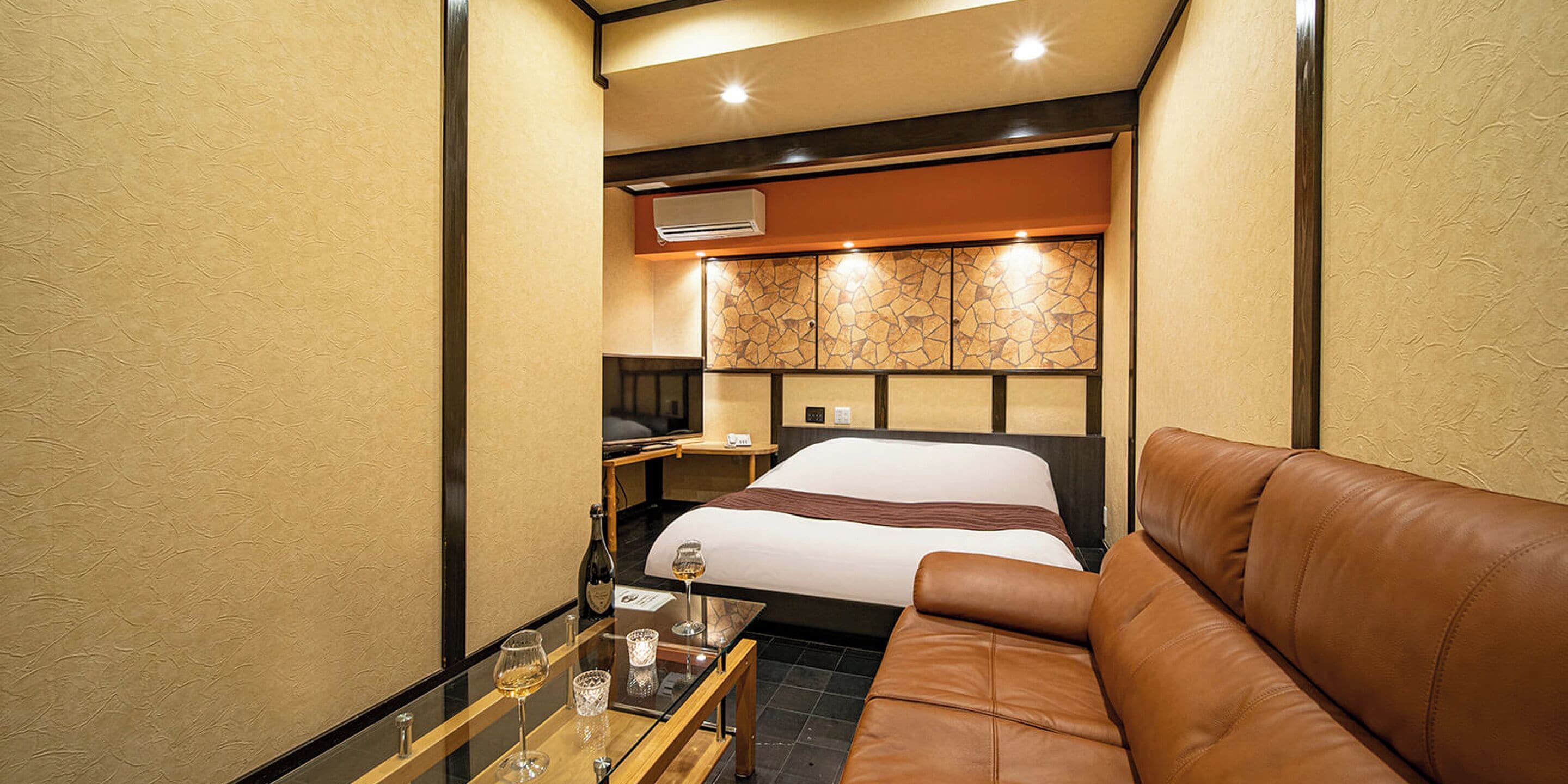 A compact hotel room with a bed, a leather sofa, a small table with glasses, and a minibar.