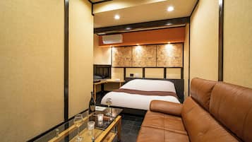 A compact hotel room with a bed, a leather sofa, a small table with glasses, and a minibar.