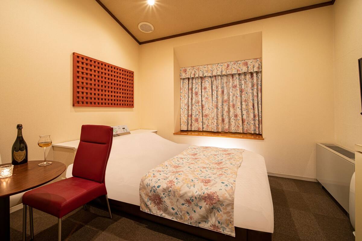 A hotel room with a bed, a red chair, a table with a bottle and glasses, a TV, and a window with curtains.