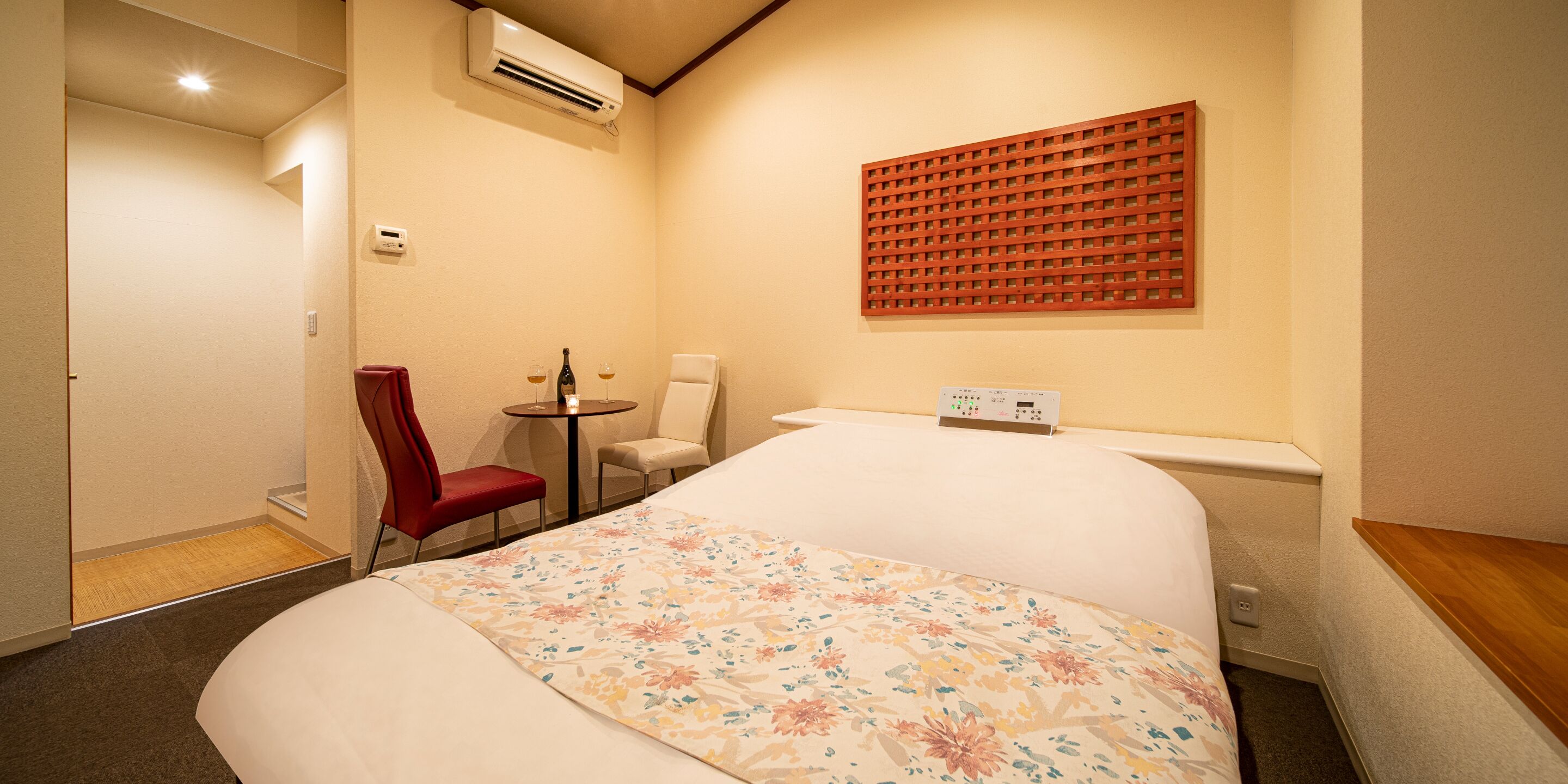 A hotel room with a large bed, a small table with two chairs, a red chair, a wall-mounted air conditioner, and a decorative panel.