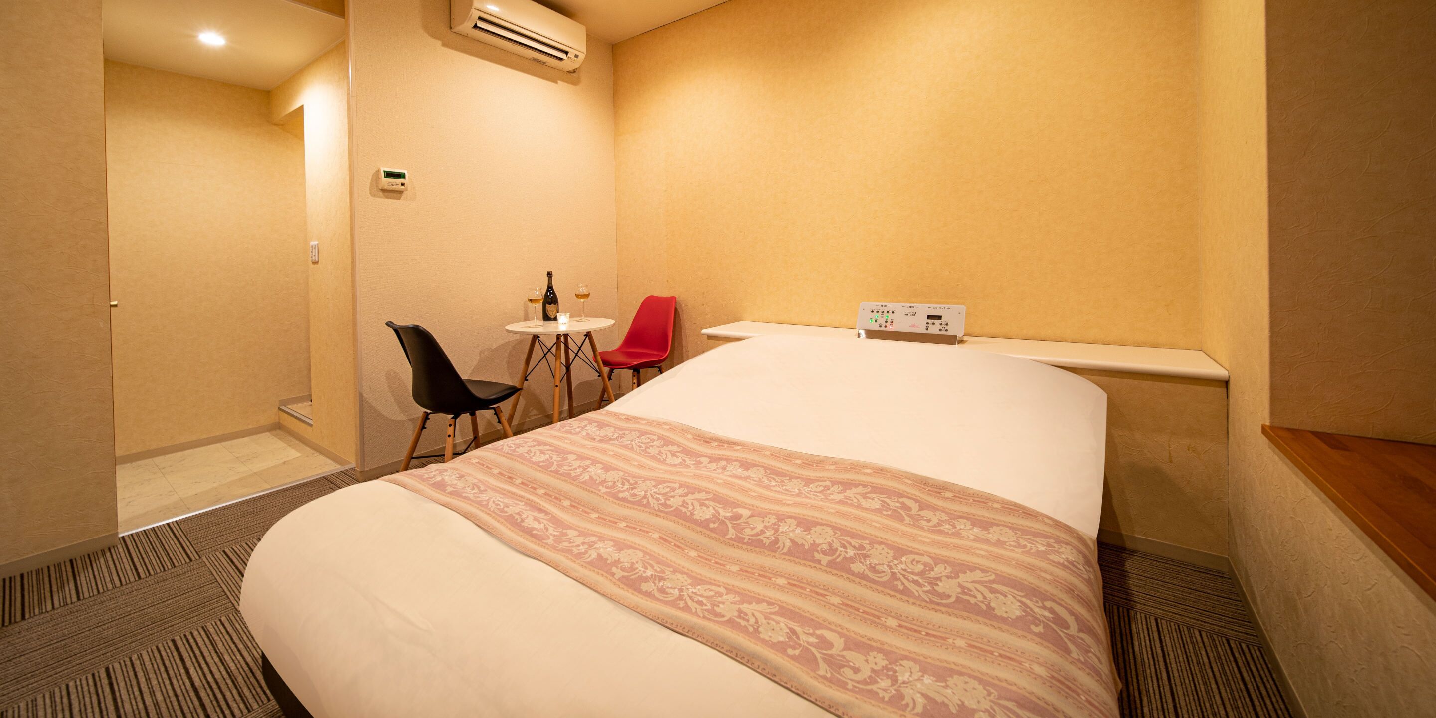 Habitación de hotel con cama, una mesita con dos sillas, un equipo de aire acondicionado y un panel de control en la pared.