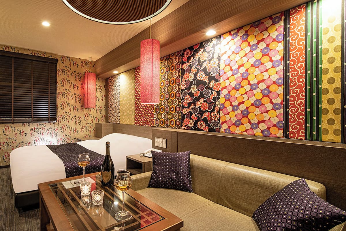 A hotel room with two beds, a sofa, a table with glasses and a bottle, and a colorful wall.