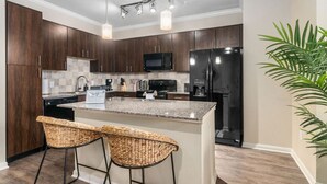 A modern kitchen with dark wood cabinets, a granite countertop, a stainless steel refrigerator, and a built-in microwave. There are two wicker bar stools and a potted plant. - Modish Keen King Bed w/ Pool (Houston)