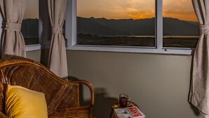 A room with a view of mountains at sunset, a wicker chair with a yellow pillow, and a table with a book and a glass of drink. - Zostel Pokhara (Pokhara)
