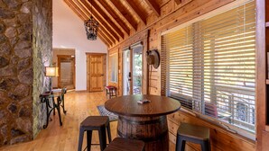 A wooden interior with a stone wall, a round wooden table with stools, and large windows with blinds.