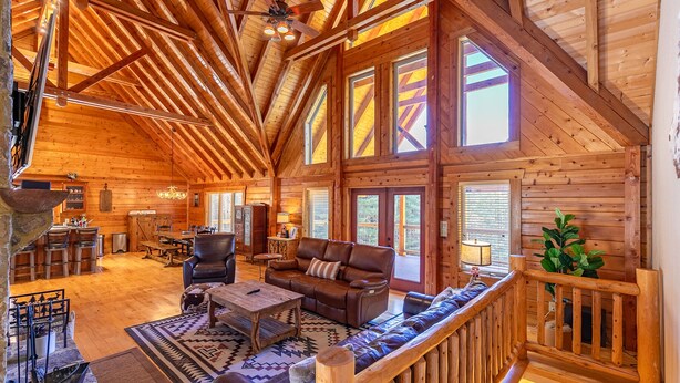 A spacious log cabin interior with a high ceiling, large windows, a cozy seating area, and a stone fireplace.