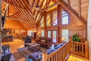 A spacious log cabin interior with a high ceiling, large windows, a cozy seating area, and a stone fireplace. - Whiskey River by the New - hot tub, game room, New River access! (Jefferson)