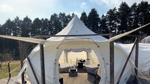 A large, white tent with a peaked roof, set on a wooden deck. Inside, there is a bed, a small table, and a bench.