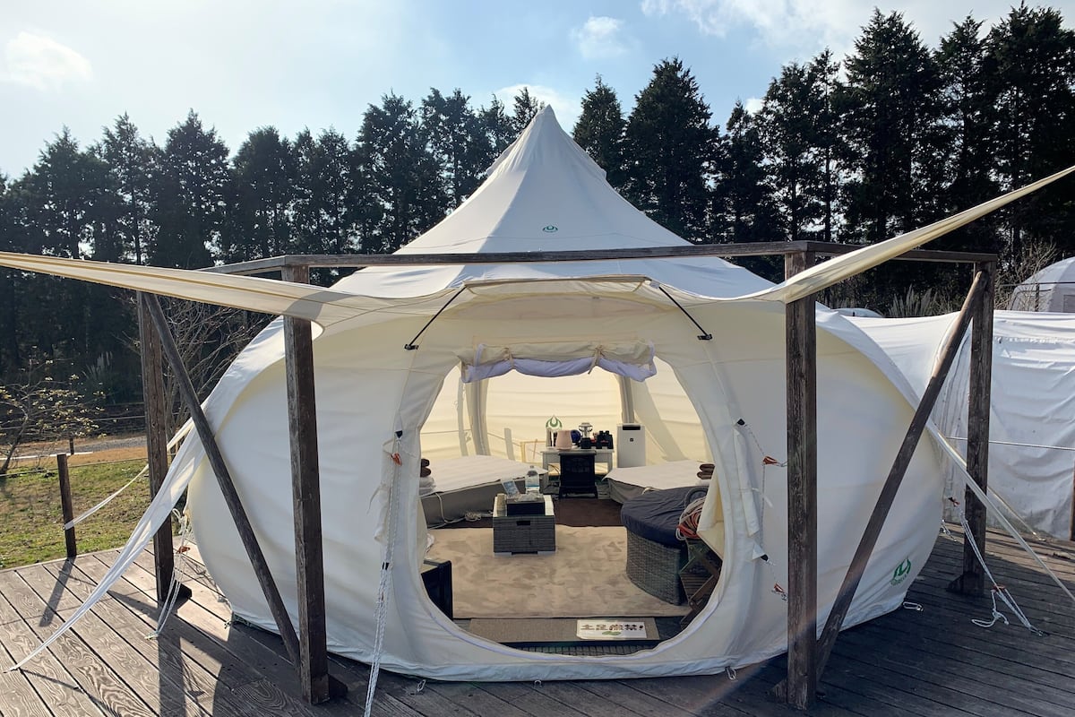 A large, white tent with a peaked roof, set on a wooden deck. Inside, there is a bed, a small table, and a bench.