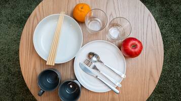 A round wooden table with plates, cutlery, chopsticks, glasses, and a fruit.