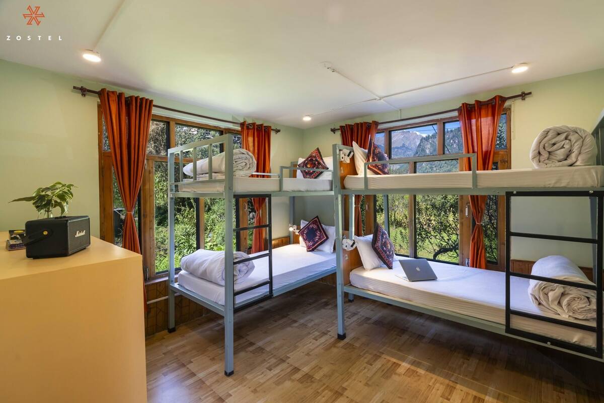 A hostel room with bunk beds, wooden floors, and large windows with curtains.