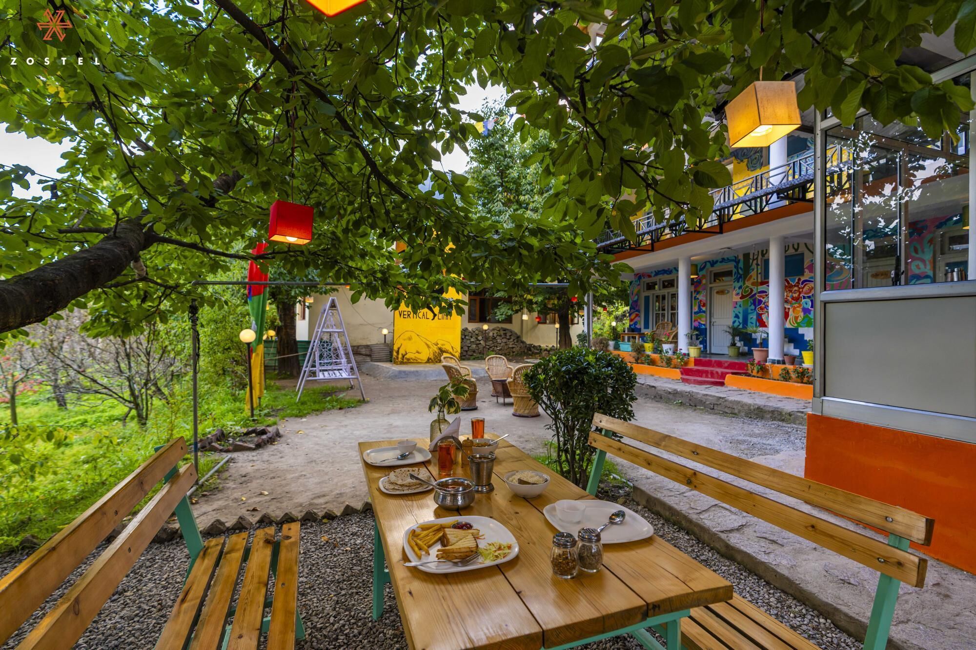 Un espace extérieur aménagé pour les repas, avec une table en bois pour deux personnes, entouré de verdure et avec un bâtiment coloré en arrière-plan.