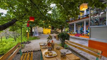 Un espace extérieur aménagé pour les repas, avec une table en bois pour deux personnes, entouré de verdure et avec un bâtiment coloré en arrière-plan.