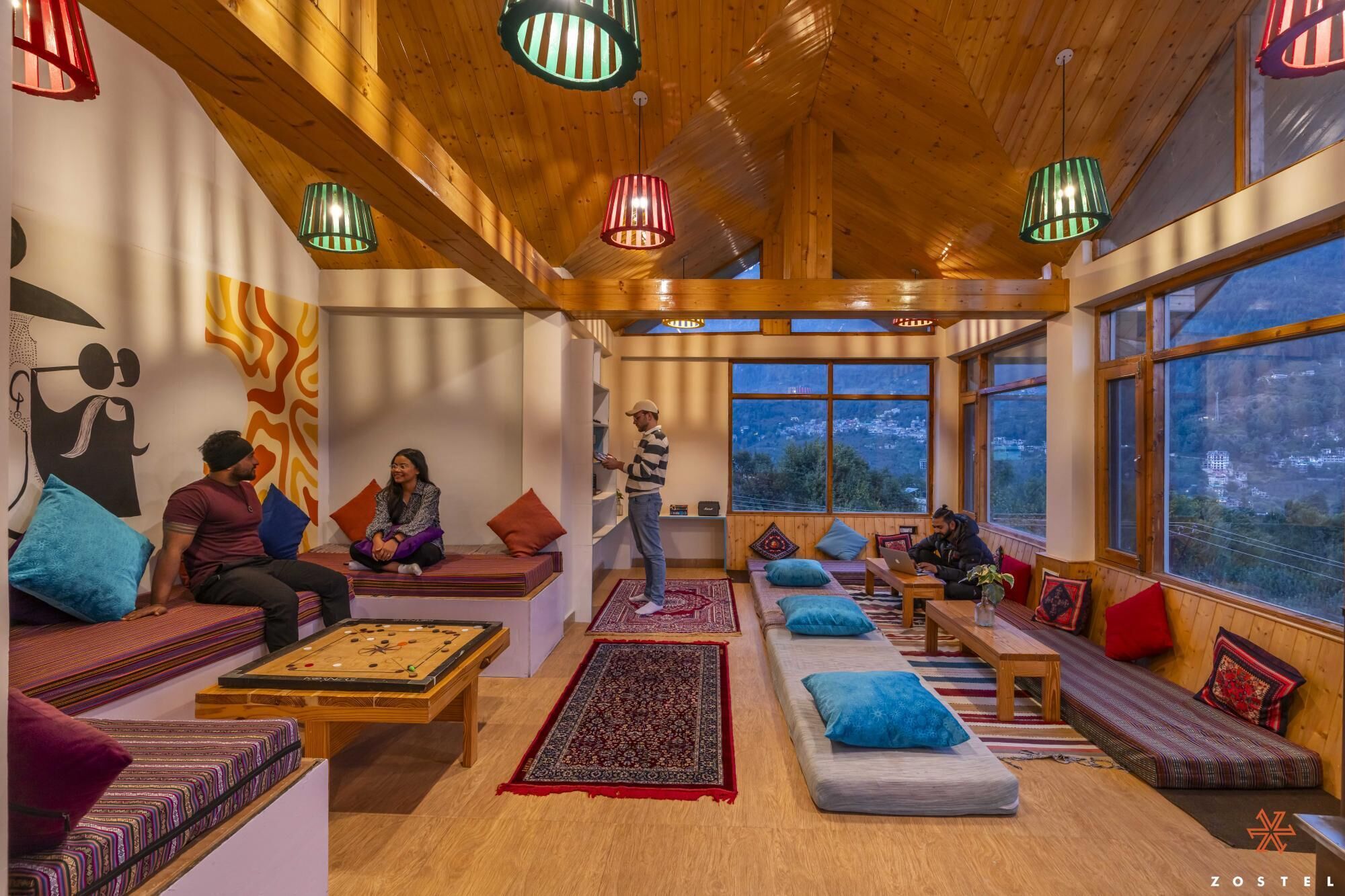 A cozy room with wooden beams, colorful cushions, a pool table, and large windows offering a view of the landscape.