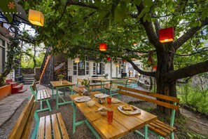 An outdoor dining area with wooden tables and benches, surrounded by trees and a building with a brick facade. - Zostel Old Manali (Manali)