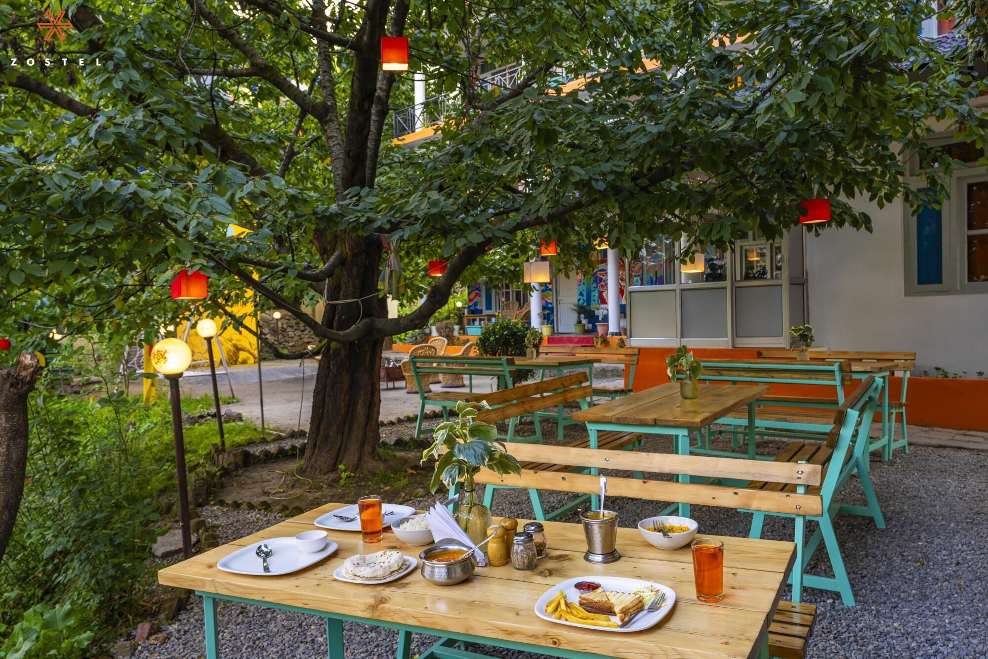 An outdoor dining area with wooden tables and benches, surrounded by greenery and trees.