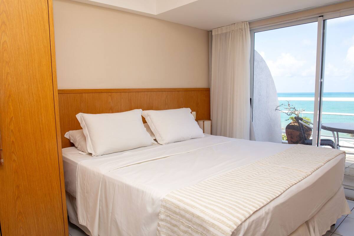 A hotel room with a large bed, wooden headboard, and a view of the ocean through a large window.