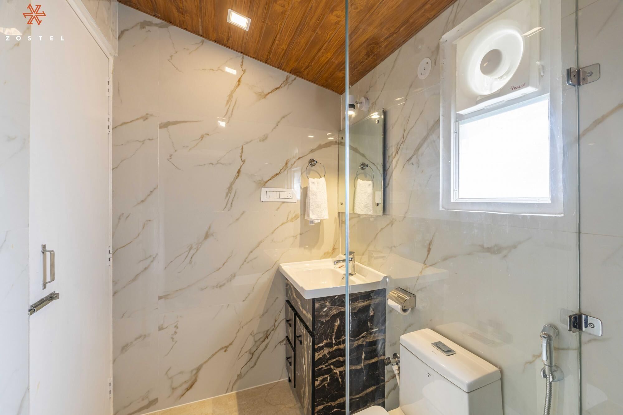 Un baño moderno con paredes de mármol, cabina de ducha con mampara de cristal, lavabo blanco y un inodoro.
