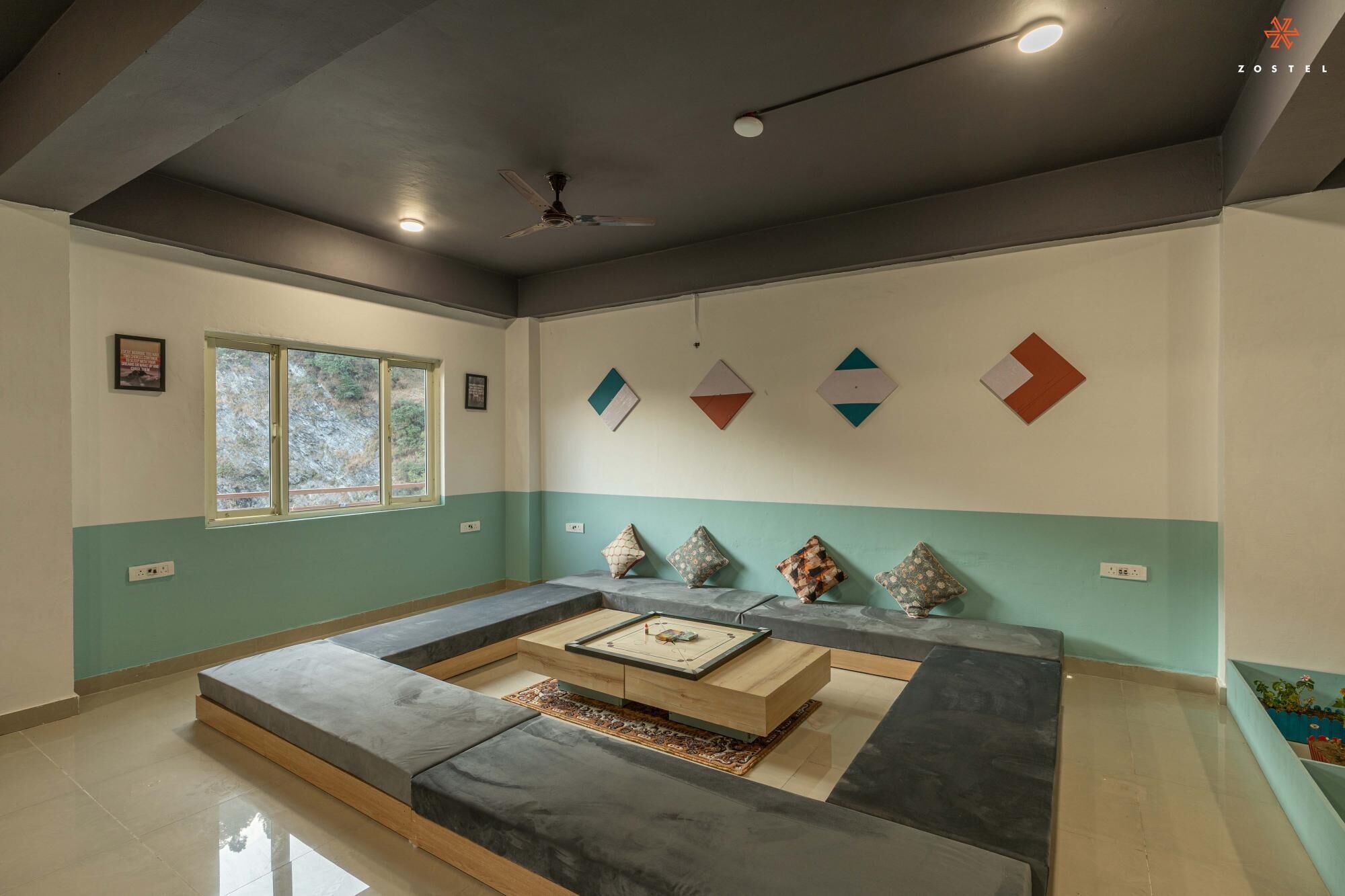 Un salon moderne avec un canapé d’angle, une table basse et des coussins décoratifs. Une fenêtre offre une vue sur les arbres, et des œuvres d’art géométriques ornent le mur.