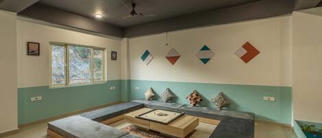 Un salon moderne avec un canapé d’angle, une table basse et des coussins décoratifs. Une fenêtre offre une vue sur les arbres, et des œuvres d’art géométriques ornent le mur.