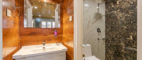 Un baño con tocador de madera, lavamanos, espejo y ducha con paredes de mármol.