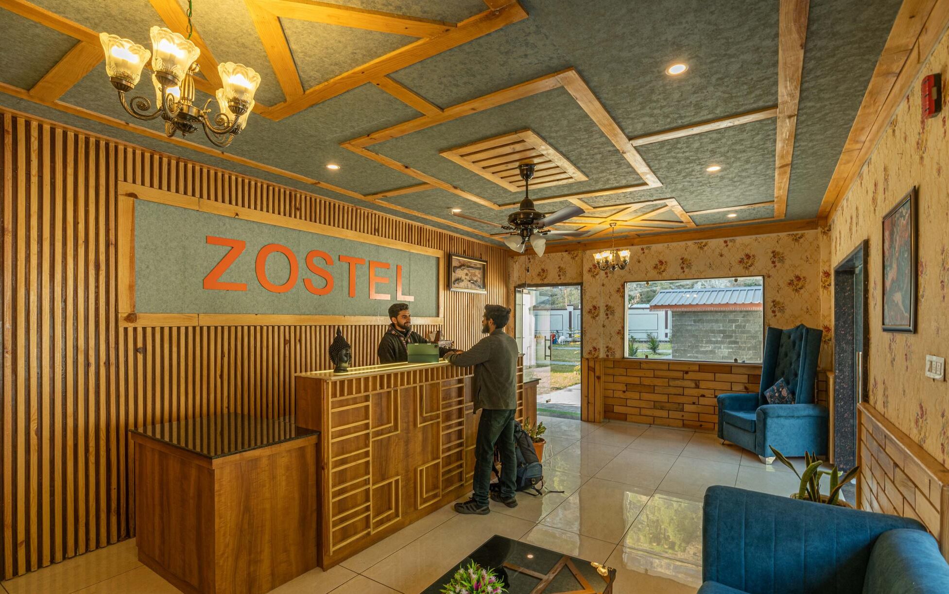 A reception area with a wooden reception desk, a sign reading "ZOSTEL", and a ceiling fan.